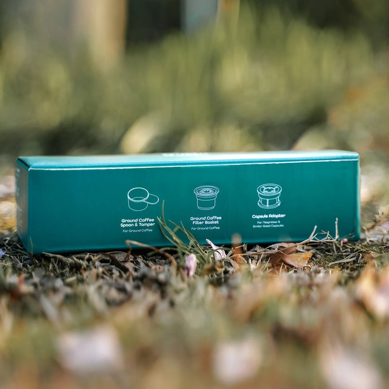 Синьо-зелена кутия Комплект адаптери Outin Nano с бели икони седи на трева, показвайки методи за приготвяне на кафе. Синьо-зелена кутия Комплект адаптери Outin Nano с бели икони седи на трева, показвайки методи за приготвяне на кафе.