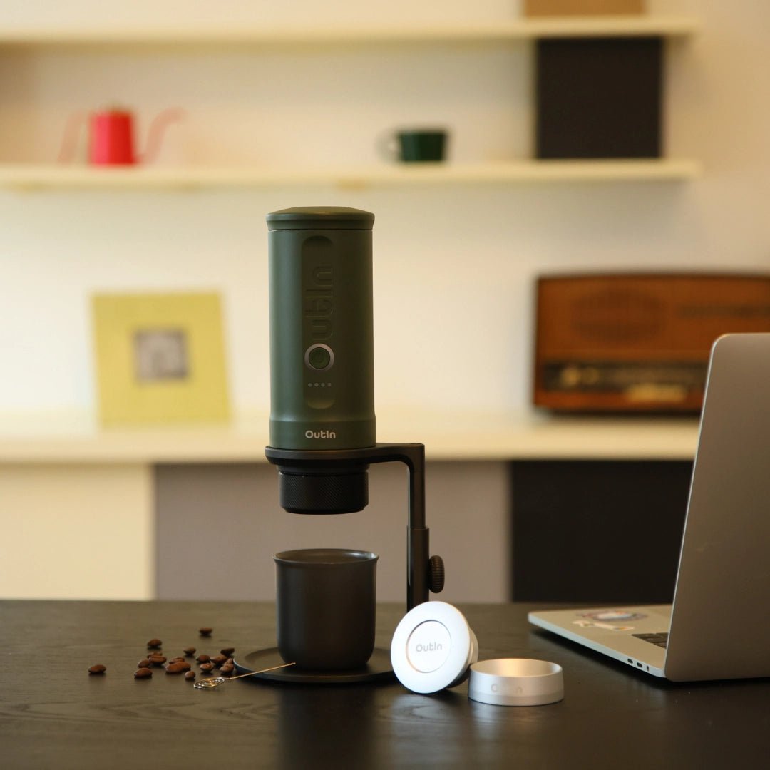 Метална стойка за Outin Nano, варяща се в чаша до лаптоп, с утайка от кафе, разпръсната наблизо.