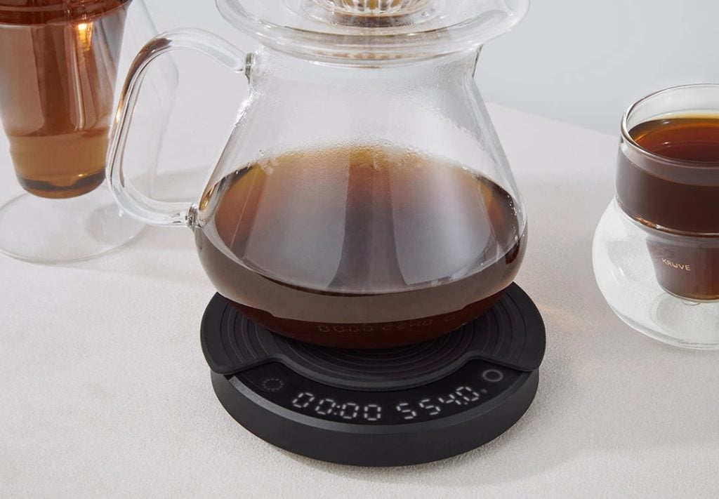 Стъклен съд за кафе Outin стои на цифрова везна, до чаша с кафе и друга празна чаша.