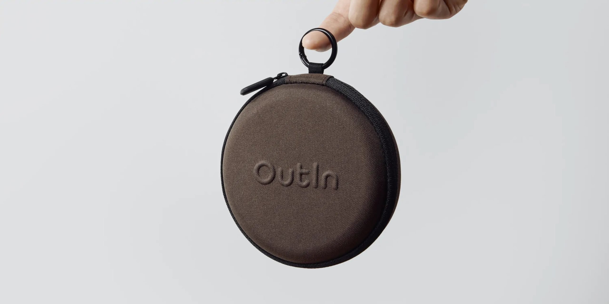 Една ръка държи кръгъл калъф за носене на Outin с цип в кафяв цвят с релефно име на марката на обикновен фон.