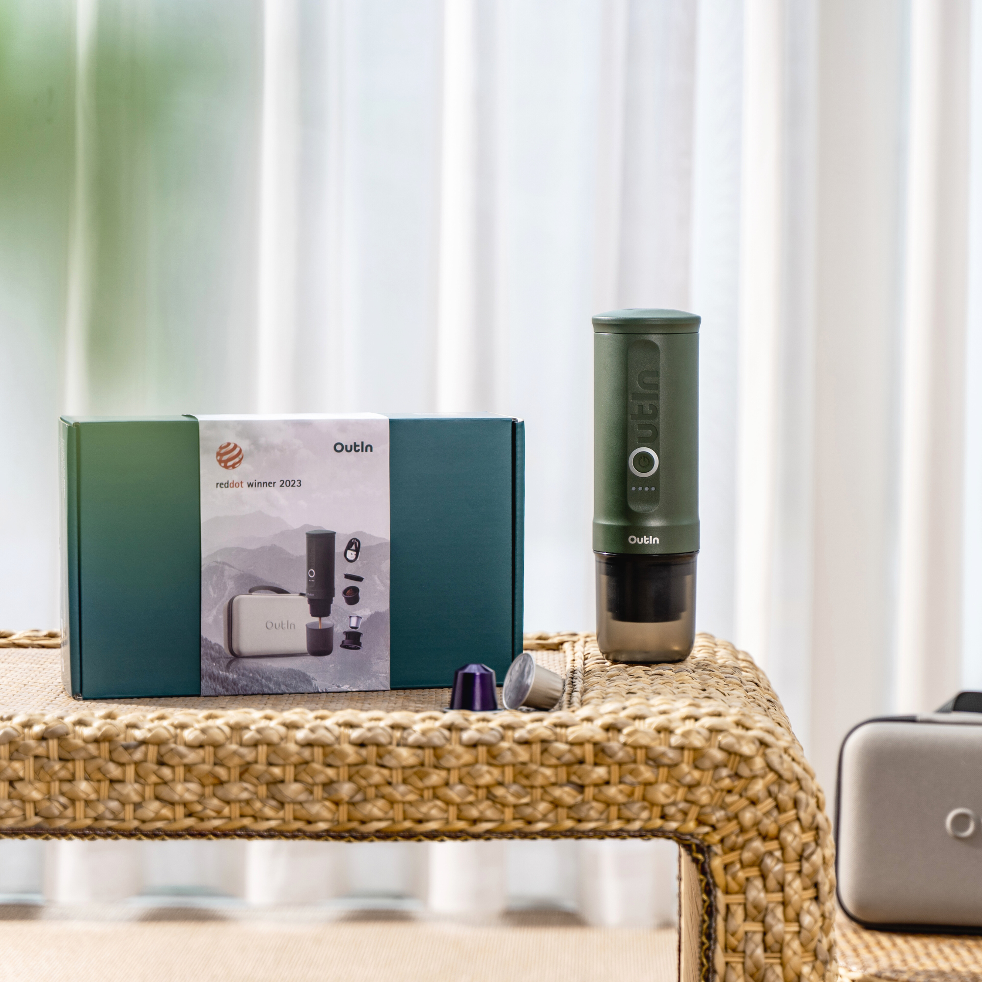 A Портативна кафемашина за еспресо Outin Nano + Калъф и кутията са изложени до кафе капсули върху тъкана маса.