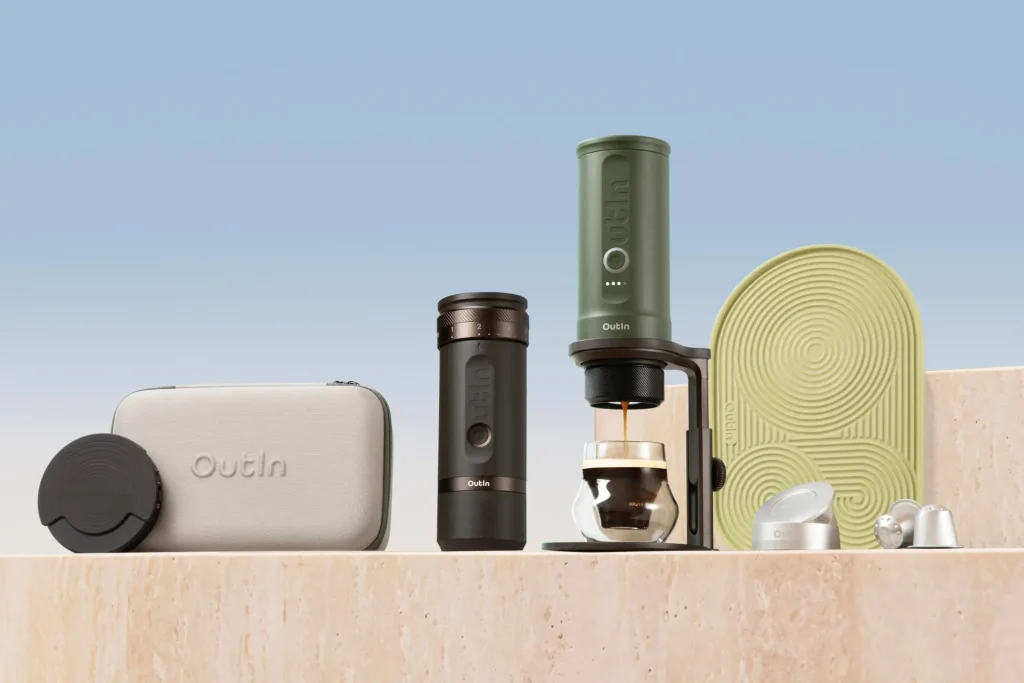 Комплект преносима кафемашина Outin с аксесоари, калъф за носене, чаша и силиконова подложка върху бежова повърхност.