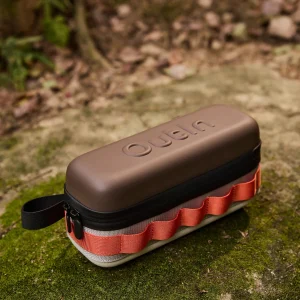 Калъф с твърда черупка Outin в кафяво и оранжево, с цип и каишка, лежи на мъхеста повърхност на открито.