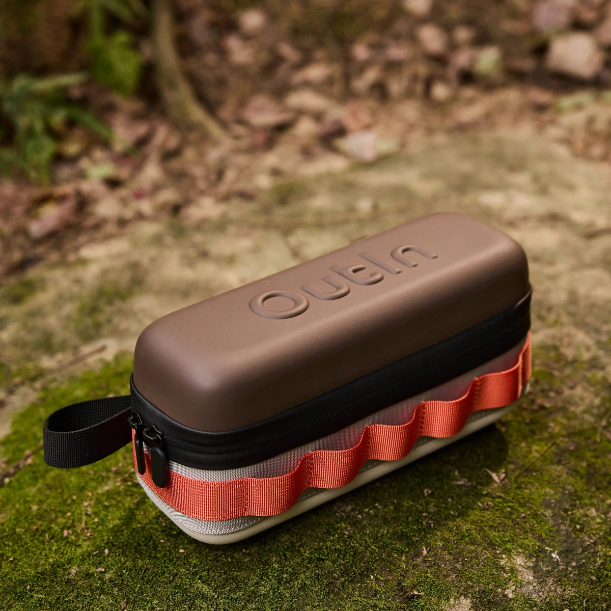 Калъф с твърда черупка Outin в кафяво и оранжево, с цип и каишка, лежи на мъхеста повърхност на открито. Калъф с твърда черупка Outin в кафяво и оранжево, с цип и каишка, лежи на мъхеста повърхност на открито.