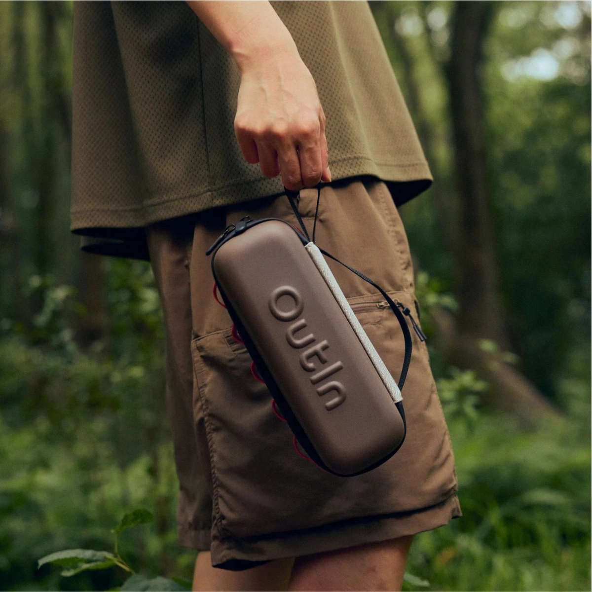 Човек с кафяви къси панталони държи преносим калъф Outin в спокойна горска обстановка. Човек с кафяви къси панталони държи преносим калъф Outin в спокойна горска обстановка.