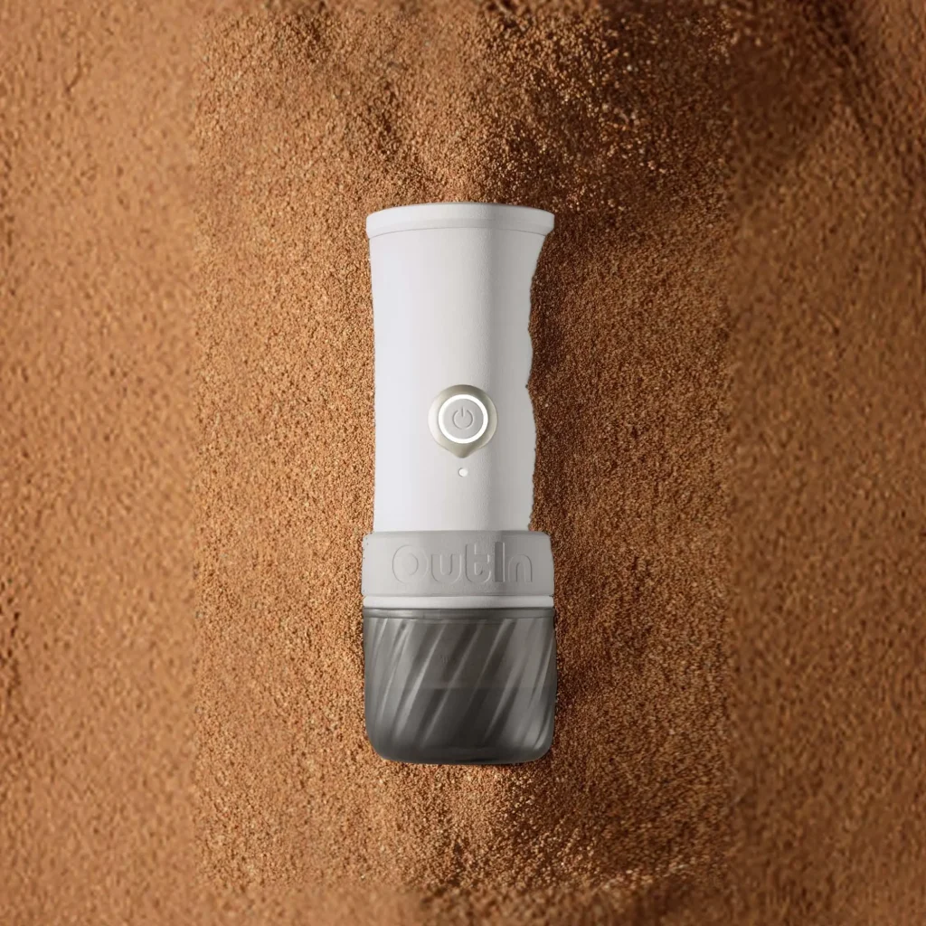 Преносима кафемелачка Outin Mino с бутон за включване лежи върху легло от смляно кафе.