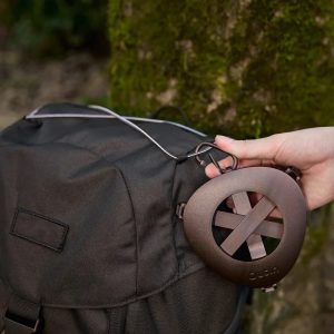 Ръка прикрепя черно триъгълно устройство към черна раница с метална щипка; на заден план - мъхесто дърво.